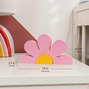 K&ouml;nnte beinhalten: Eine rosa Holzblumen-Dekoration mit gelber Mitte. Die Blume ist 19,3 cm breit, 11,4 cm hoch und 3 cm dick. Der Dekorationsartikel befindet sich auf einer wei&szlig;en Oberfl&auml;che.