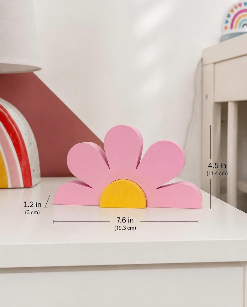 K&ouml;nnte beinhalten: Eine rosa Holzblumen-Dekoration mit gelber Mitte. Die Blume ist 19,3 cm breit, 11,4 cm hoch und 3 cm dick. Der Dekorationsartikel befindet sich auf einer wei&szlig;en Oberfl&auml;che.