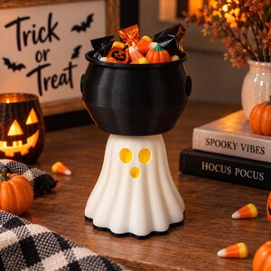 May include: A black cauldron bowl resting on a white ghost-shaped base with three cut-out holes for eyes and a mouth. The cauldron is filled with assorted Halloween sweets, including orange pumpkin-shaped treats and black-wrapped items.