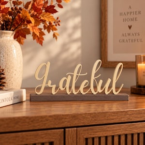 May include: A decorative wooden sign featuring the word Grateful in a cursive script font, mounted on a rectangular dark gray base. The sign is crafted from light-colored wood and stands upright, displaying the text clearly in a single line.