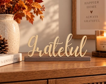 Puede incluir: Un letrero decorativo de madera con la palabra Grateful en una fuente cursiva, montado sobre una base rectangular de color gris oscuro. El letrero está hecho de madera clara y se mantiene en posición vertical, mostrando el texto en una línea.