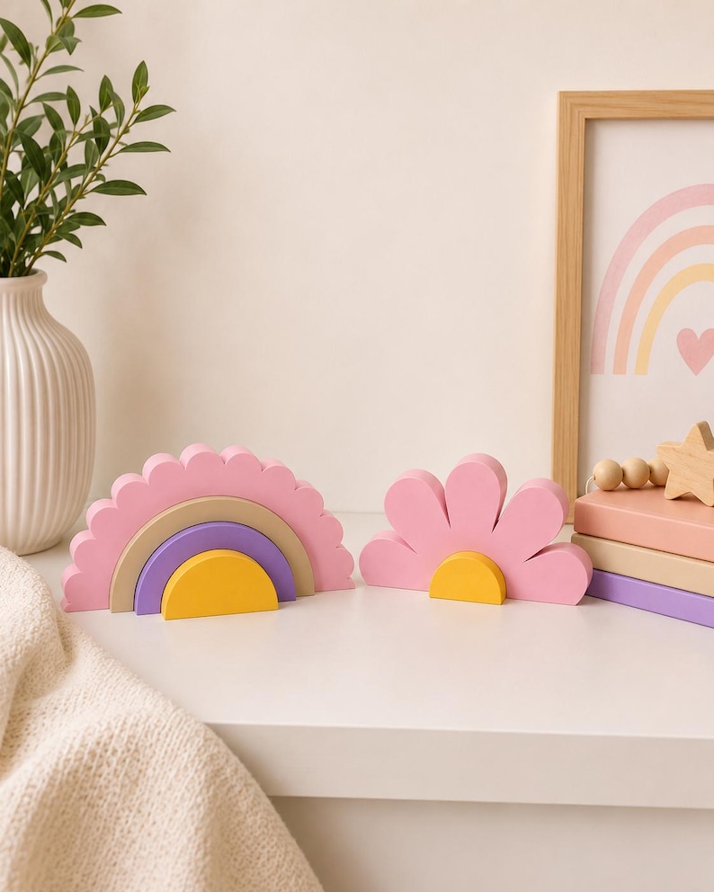 K&ouml;nnte beinhalten: Zwei dekorative Holzst&uuml;cke: ein rosa Regenbogen mit gelben, lila und beigen Schichten und eine rosa Blume mit gelber Mitte. Sie stehen auf einer wei&szlig;en Oberfl&auml;che, mit einem gerahmten Regenbogen-Druck im Hintergrund. Eine wei&szlig;e Vase mit Gr&uuml;nzeug befindet sich links.