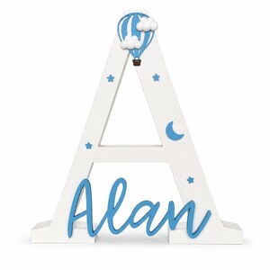 May include: A white wooden letter "A" with blue accents. The letter is decorated with a blue hot air balloon, clouds, stars, and a crescent moon. The name "Alan" is written in blue script across the base.