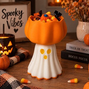 May include: A white ghost-shaped bowl stand with a bright orange pumpkin-shaped bowl on top, filled with assorted Halloween sweets including small pumpkins and wrapped treats. The base features three cut-out holes for eyes and a mouth.