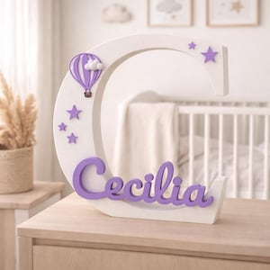 May include: A white wooden letter "C" with the name "Cecilia" in purple script. The letter is decorated with purple stars and a hot air balloon. A decorative item for a nursery or child's room.