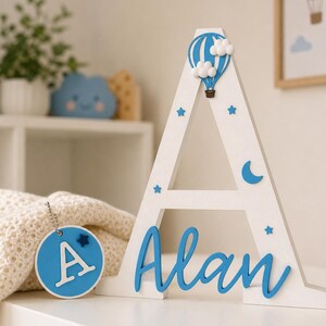 May include: A white wooden letter "A" with blue accents, including a hot air balloon, stars, and the name "Alan" in cursive. A blue circular tag with the letter "A" and a star hangs nearby. Decorative nursery decor.