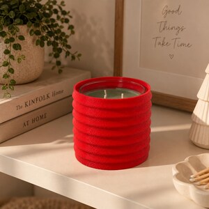 Puede incluir: Una vela cil&iacute;ndrica en un recipiente de color rojo brillante, texturizado y acanalado. La vela presenta una superficie de cera verde claro con tres mechas visibles. El exterior del recipiente tiene un acabado mate y t&aacute;ctil con crestas horizontales.