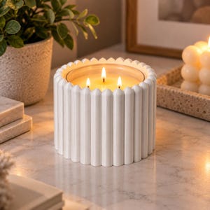 May include: A white cylindrical candle holder with a vertical ribbed texture containing a three-wick candle with a glowing flame. The candle is positioned on a light-colored surface, emitting a warm light from the three lit wicks inside the vessel.