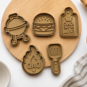 May include: A set of five brown plastic cookie cutters featuring a grill, a hamburger, a rectangular sign with the text KING OF THE GRILL, a flame shape with the text DAD, and a spatula. The cutters are arranged on a round wooden board.