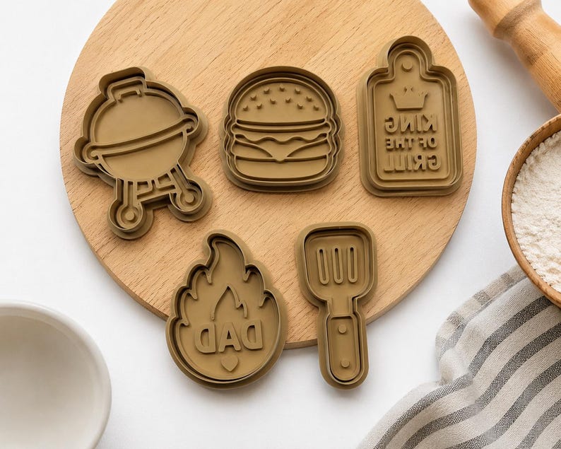 May include: A set of five brown plastic cookie cutters featuring a grill, a hamburger, a rectangular sign with the text KING OF THE GRILL, a flame shape with the text DAD, and a spatula. The cutters are arranged on a round wooden board.
