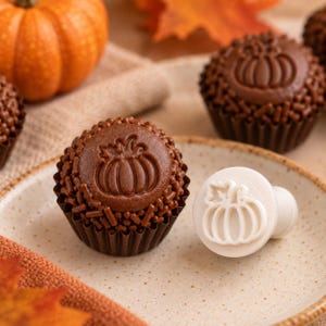 May include: A round chocolate truffle coated in brown sprinkles, featuring a stamped pumpkin design on top, placed in a dark brown paper liner next to a white plastic stamp tool with a matching pumpkin motif on a speckled plate.