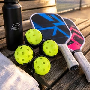 Puede incluir: Una botella de agua negra con un logotipo S blanco, cuatro pelotas de pickleball de color amarillo ne&oacute;n perforadas en un soporte negro, dos palas de pickleball con empu&ntilde;aduras blancas y patrones geom&eacute;tricos azules o rosas, y una toalla blanca doblada.