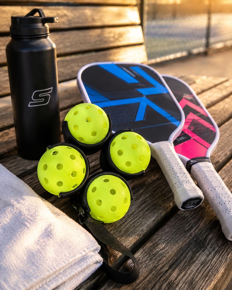 Puede incluir: Una botella de agua negra con un logotipo S blanco, cuatro pelotas de pickleball de color amarillo ne&oacute;n perforadas en un soporte negro, dos palas de pickleball con empu&ntilde;aduras blancas y patrones geom&eacute;tricos azules o rosas, y una toalla blanca doblada.