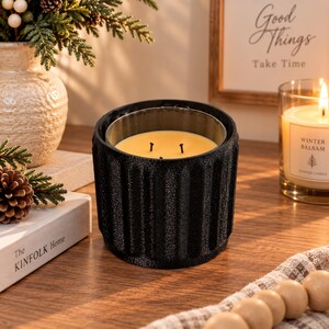 May include: A black, cylindrical candle holder with a vertical ribbed texture and a matte finish. The holder contains a glass insert filled with yellow wax and features three black wicks positioned in the centre of the wax surface.