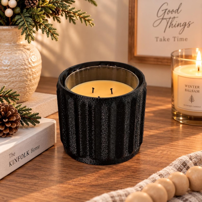 May include: A black, cylindrical candle holder with a vertical ribbed texture and a matte finish. The holder contains a glass insert filled with yellow wax and features three black wicks positioned in the centre of the wax surface.