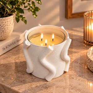 May include: A white three-wick candle in a cylindrical container with a textured surface featuring raised, wavy, snake-like decorative elements. The candle wax is light yellow, and the three wicks are lit, producing small, bright, flickering flames.