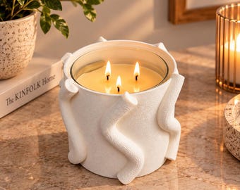 Puede incluir: Una vela blanca de tres mechas en un recipiente cilíndrico con una superficie texturizada que presenta elementos decorativos en relieve, ondulados y con forma de serpiente. La cera es de color amarillo claro y las tres mechas están encendidas.