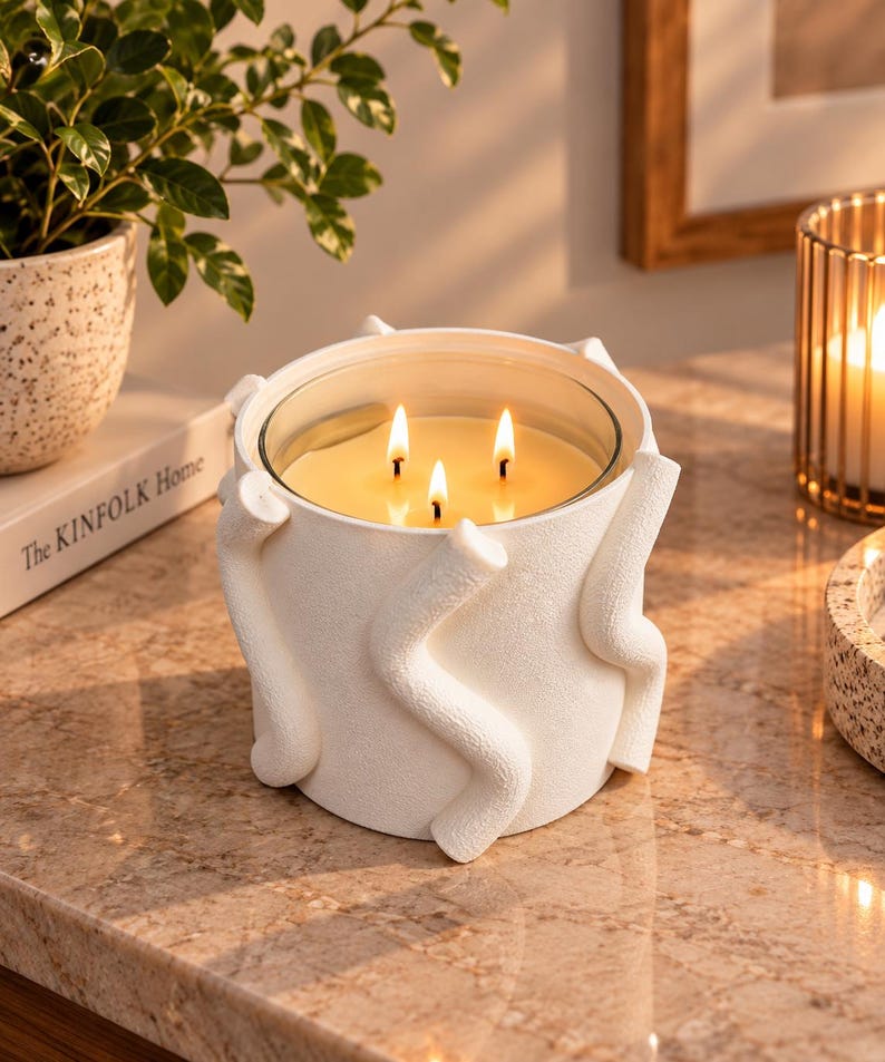 May include: A white three-wick candle in a cylindrical container with a textured surface featuring raised, wavy, snake-like decorative elements. The candle wax is light yellow, and the three wicks are lit, producing small, bright, flickering flames.