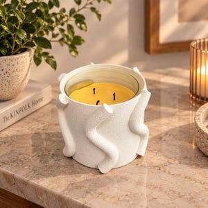 May include: A three-wick candle in a white cylindrical vessel with a textured surface featuring raised, wavy, vertical lines. The wax inside is yellow, and the candle is contained within a clear glass insert set inside the decorative white holder.