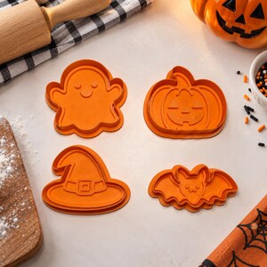 May include: A set of four orange plastic cookie cutters featuring Halloween-themed shapes: a ghost with a smiling face, a jack-o'-lantern pumpkin, a pointed witch hat with a buckle detail, and a bat with wings spread and a small smiling face.