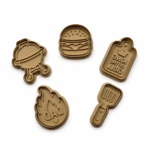 May include: A set of five tan plastic cookie cutters featuring barbecue-themed shapes including a round grill, a hamburger, a rectangular plaque with the text "KING OF THE GRILL", a flame shape with the word "DAD", and a slotted spatula tool.