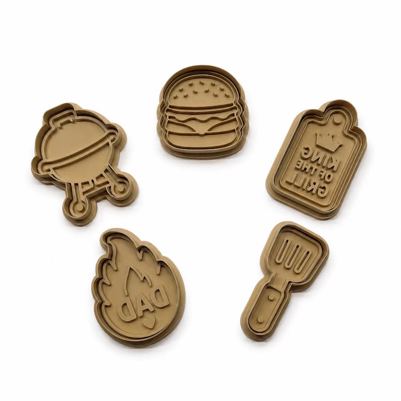 May include: A set of five tan plastic cookie cutters featuring barbecue-themed shapes including a round grill, a hamburger, a rectangular plaque with the text "KING OF THE GRILL", a flame shape with the word "DAD", and a slotted spatula tool.