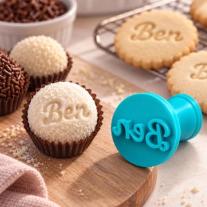 May include: A bright turquoise plastic stamp with the name Ben in cursive script, positioned next to a round white confection in a brown paper liner that is embossed with the same name Ben in a matching cursive script style.