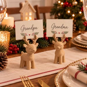 May include: Two light brown reindeer-shaped place card holders with textured surfaces. Each holds a rectangular white card with a green leaf and red berry design. One card features the text "Grandpa" and the other features the text "Grandma".