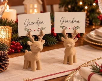 May include: Two light brown reindeer-shaped place card holders with textured surfaces. Each holds a rectangular white card with a green leaf and red berry design. One card features the text "Grandpa" and the other features the text "Grandma".