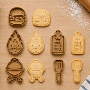 May include: A set of brown plastic cookie cutters and matching light tan sugar cookies shaped like a burger, a flame with the text DAD, a rectangular board with the text KING OF THE GRILL, a round charcoal grill, and a spatula with a handle.