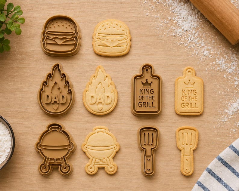 May include: A set of brown plastic cookie cutters and matching light tan sugar cookies shaped like a burger, a flame with the text DAD, a rectangular board with the text KING OF THE GRILL, a round charcoal grill, and a spatula with a handle.