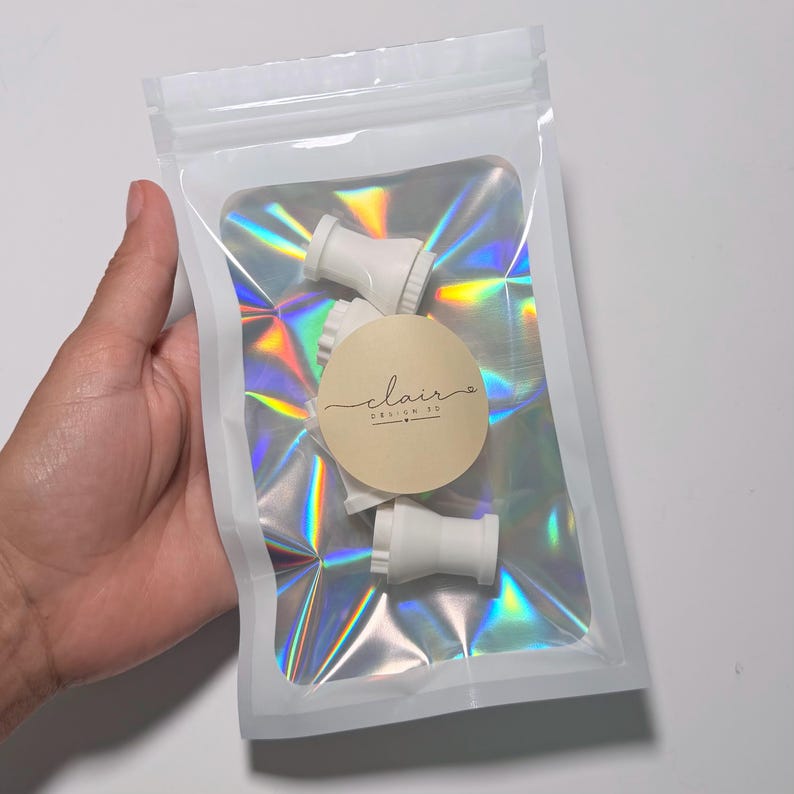 May include: A clear, holographic, resealable plastic pouch containing several small, white, cylindrical plastic components. A circular beige sticker is affixed to the front of the pouch, featuring the text "clair DESIGN CO" in a dark script font.