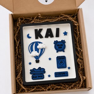 May include: A rectangular shadow box frame with a white background and black border containing blue and white 3D icons. The name KAI is at the top. Icons include a hot air balloon, alarm clock, calendar, baby silhouette, and text reading 8LB 11OZ and 19.7INCHES.