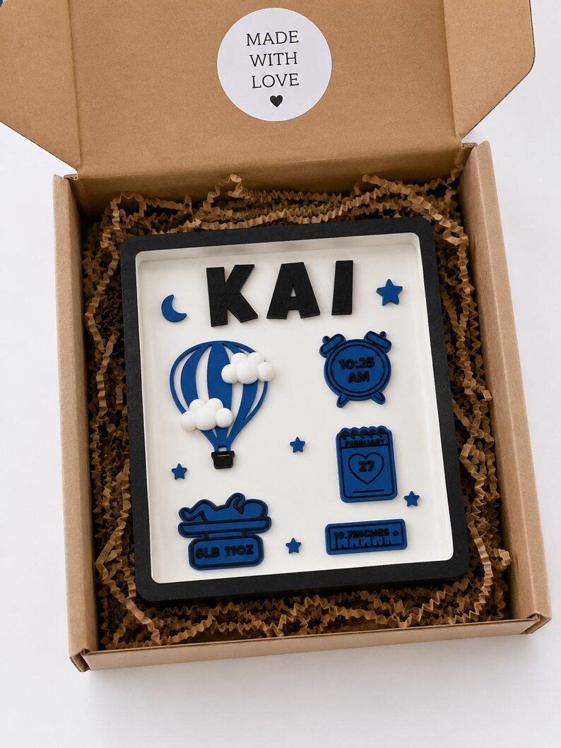 May include: A rectangular shadow box frame with a white background and black border containing blue and white 3D icons. The name KAI is at the top. Icons include a hot air balloon, alarm clock, calendar, baby silhouette, and text reading 8LB 11OZ and 19.7INCHES.