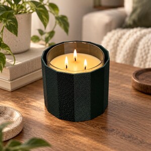Puede incluir: Una vela de tres mechas en un soporte cil&iacute;ndrico de color verde oscuro, facetado y texturizado. La cera es de color crema y las tres mechas est&aacute;n encendidas, produciendo un brillo c&aacute;lido. El recipiente tiene un dise&ntilde;o geom&eacute;trico.