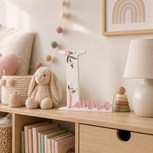 May include: A white wooden letter "I" adorned with pink flowers and green leaves. The name "Imma" is written in pink script below the letter. The letter is displayed on a wooden shelf with a stuffed bunny and other decorative items.