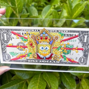 Può includere: Una banconota da un dollaro con un'illustrazione colorata di Spongebob Squarepants che indossa una corona e un mantello reale, seduto su un trono circondato da monete d'oro e banconote verdi, con linee rosse che attraversano il centro.