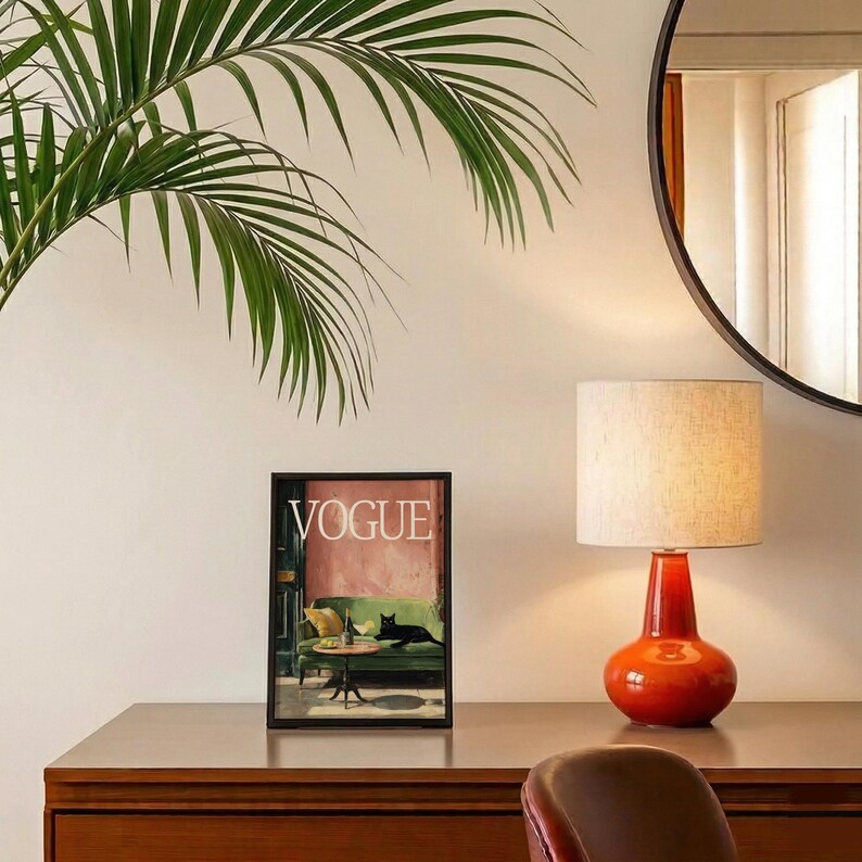 Puede incluir: Una portada enmarcada de la revista Vogue con una escena de sal&oacute;n con un gato negro, una l&aacute;mpara roja con una pantalla beige y un espejo redondo. La escena est&aacute; sobre una superficie de madera con una hoja de palma verde.