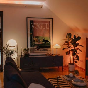 Puede incluir: Un sal&oacute;n con un gran cuadro de "VOGUE", un sof&aacute; gris oscuro y una alfombra a rayas blancas y negras. Dos l&aacute;mparas de globo y una peque&ntilde;a l&aacute;mpara de mesa iluminan el espacio. Una planta en maceta y un mueble gris oscuro completan la decoraci&oacute;n.