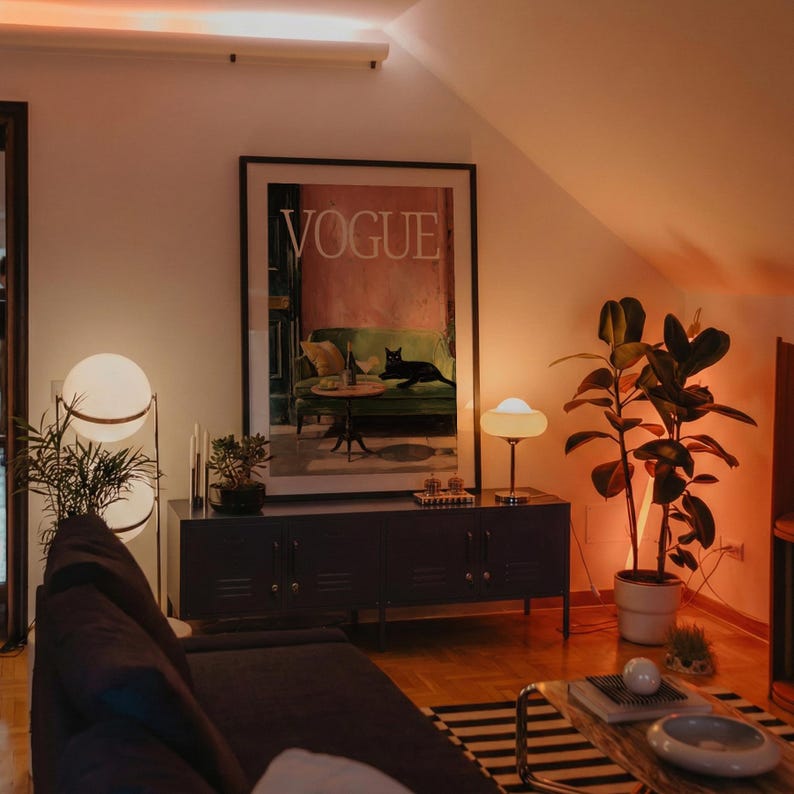 Puede incluir: Un sal&oacute;n con un gran cuadro de "VOGUE", un sof&aacute; gris oscuro y una alfombra a rayas blancas y negras. Dos l&aacute;mparas de globo y una peque&ntilde;a l&aacute;mpara de mesa iluminan el espacio. Una planta en maceta y un mueble gris oscuro completan la decoraci&oacute;n.