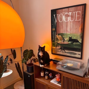 Puede incluir: Un gato blanco y negro est&aacute; sentado encima de un mueble de madera, junto a un tocadiscos y una impresi&oacute;n enmarcada de "Vogue". La habitaci&oacute;n cuenta con una gran l&aacute;mpara naranja, plantas en macetas y botellas decorativas. Tambi&eacute;n se ve una peque&ntilde;a figura de Peppa Pig.