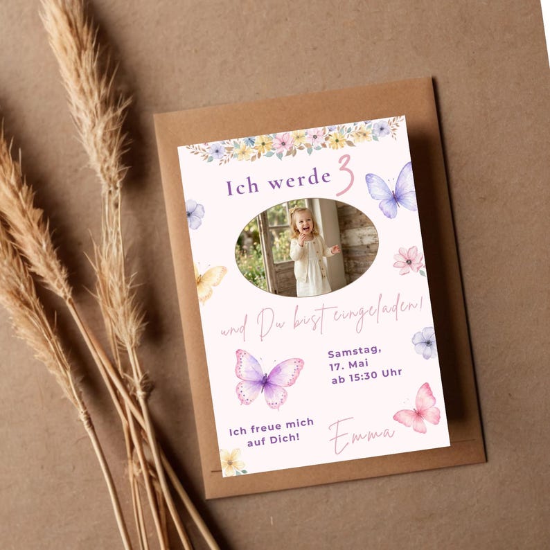 May include: A rectangular birthday invitation card featuring a central oval photo of a child, decorated with watercolor butterflies and flowers in shades of pink, purple, and yellow. The text reads Ich werde 3, und Du bist eingeladen, Samstag, 17. Mai ab 15:30 Uhr, Ich freue mich auf Dich, and Emma.