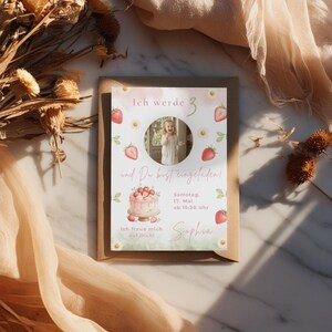 May include: A rectangular birthday invitation card featuring a circular photo of a child, watercolor strawberries, daisies, and a cake illustration. The text reads: Ich werde 3, und Du bist eingeladen!, Samstag, 17. Mai ab 15:30 Uhr, Sophia, Ich freue mich auf Dich!