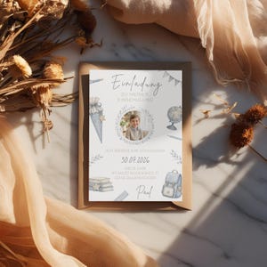 Pu&ograve; includere: Un biglietto d'invito con il testo "Einladung zu meiner Einschulung" e la foto di un bambino. La carta presenta illustrazioni di oggetti scolastici come uno zaino, libri e un mappamondo. &Egrave; visibile anche la data 30.07.2026.