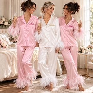 May include: Three satin pajama sets featuring long-sleeved button-down shirts and matching trousers. Two sets are light pink with the names Olivia and Sophia embroidered in pink, while the center set is white with Bride embroidered in gold.