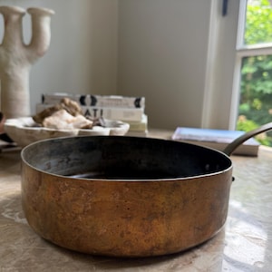 May include: A copper-colored saucepan with a long, thin handle. The pan has a dark interior and sits on a marble surface. The pan is round and has a slightly textured surface. The handle is a lighter color than the pan.