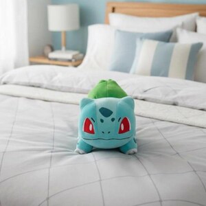 K&ouml;nnte beinhalten: Ein hellblaues Bulbasaur-Pl&uuml;schtier mit gr&uuml;nem Knollen auf dem R&uuml;cken und roten Augen liegt auf einem Bett. Das Bett hat wei&szlig;e Bettw&auml;sche und hellblaue Kissen. Der Raum hat eine hellblaue Wand.
