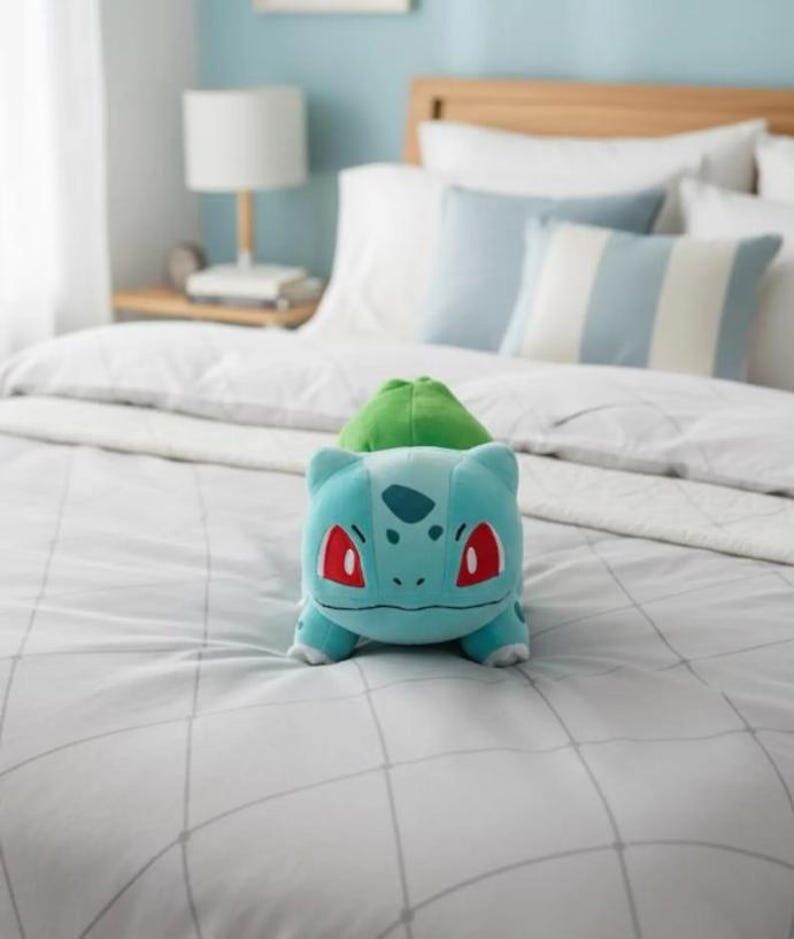 K&ouml;nnte beinhalten: Ein hellblaues Bulbasaur-Pl&uuml;schtier mit gr&uuml;nem Knollen auf dem R&uuml;cken und roten Augen liegt auf einem Bett. Das Bett hat wei&szlig;e Bettw&auml;sche und hellblaue Kissen. Der Raum hat eine hellblaue Wand.