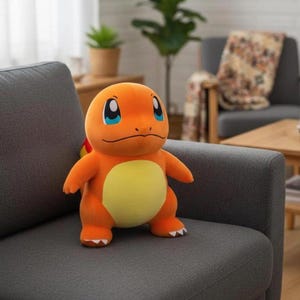 K&ouml;nnte beinhalten: Ein orangefarbenes Charmander-Pl&uuml;schtier mit gelbem Bauch und blauen Augen sitzt auf einem grauen Sofa. Das Spielzeug hat eine kleine Flamme am Schwanz. Wohnzimmer-Atmosph&auml;re im Hintergrund.