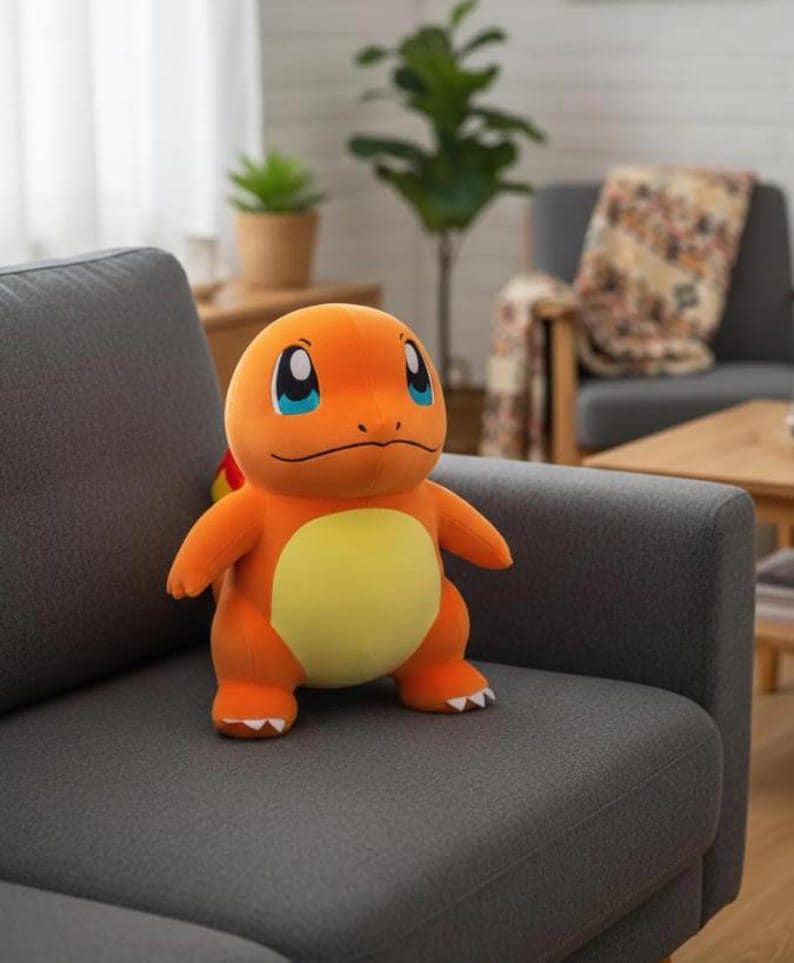 K&ouml;nnte beinhalten: Ein orangefarbenes Charmander-Pl&uuml;schtier mit gelbem Bauch und blauen Augen sitzt auf einem grauen Sofa. Das Spielzeug hat eine kleine Flamme am Schwanz. Wohnzimmer-Atmosph&auml;re im Hintergrund.
